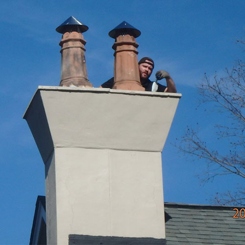 Crown Coat Application - customized chimney that flares at the top with two decorative caps.  The tech in applying crown coat.  There is a bright blue sky in the background.