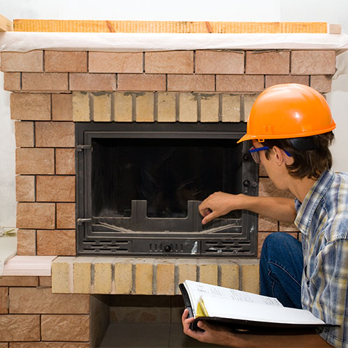 Tech wearing an orange hard hat with blue checkered shirt holding a notebook inspecting a firebox.  The surround of the firebox is terra cotta and cream.