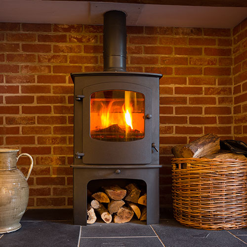 Black Wood Burning Stove with fire.  It has wood storage underneath and it is sitting in a red brick alcove.  It has a large basket of wood to the right and an urn to the left.