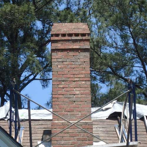Masonry Repair - Tall chimney with decorative top - scaffolding around chimney and tarps on the roof.  There are trees and blue sky in the background.